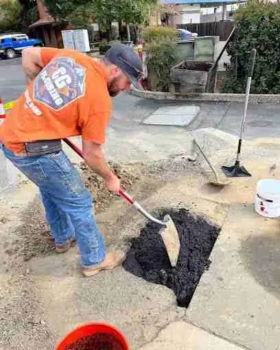 Construction worker in safety vest and hard hat repairing a pothole on a residential road in Sacramento County, California. The worker is using asphalt to fill and level the damaged pavement.