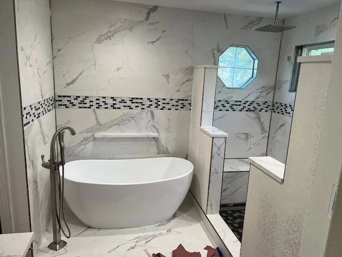 Modern bathroom with white marble tiles, a freestanding oval bathtub, black and white mosaic tile accent, walk-in shower with rain showerhead—expertly finished by a top plumbing contractor Sacramento County, CA. Octagonal window lets in natural light.