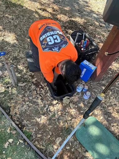 A worker in an orange "GG Plumbing" shirt inspects an open utility box outdoors, surrounded by fallen leaves and plumbing tools—a reliable choice for plumber services El Dorado, CA or a trusted plumbing contractor Sacramento County.