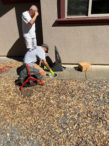 Two men, likely from a plumbing contractor in Sacramento County, CA, work outside near a building wall; one kneels to check or fix a pipe opening while the other shades his eyes. Tools and a cardboard box are on the ground nearby.