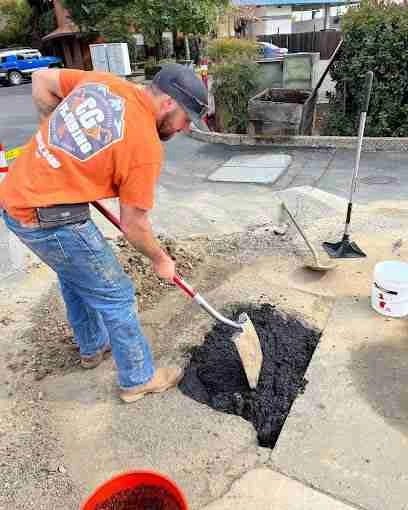 A man in an orange shirt uses a shovel to fill a hole in the pavement with asphalt on a street in Sacramento County, CA. Construction tools and a white bucket are nearby, suggesting the work of a local plumbing contractor.