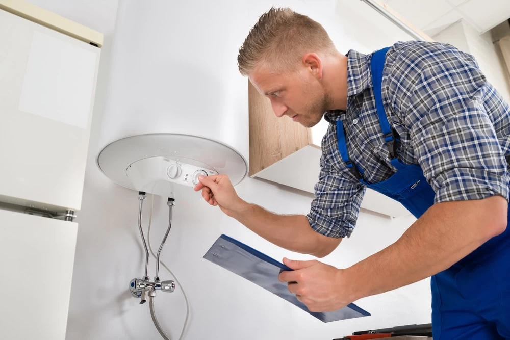 A man in blue overalls adjusts the settings on a wall-mounted water heater while holding a clipboard, appearing to inspect or repair the appliance in a modern interior.