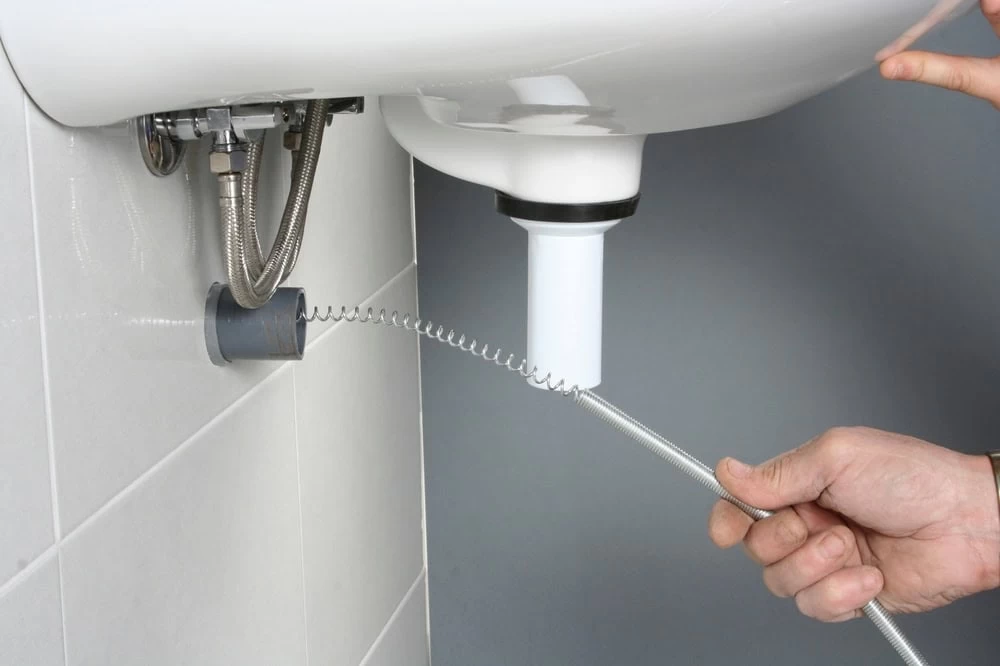 A person uses a plumber’s snake to clear a clog in a white sink drain pipe, with the snake inserted into the pipe under the sink in El Dorado, CA