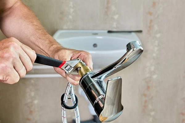 A person uses a wrench to tighten a fitting on a chrome bathroom faucet, showcasing the attention to detail provided by plumber services El Dorado,CA. Visible hoses and a sink are in the background.