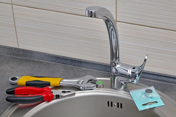 A kitchen sink with a chrome faucet is shown. Nearby, pliers, an adjustable wrench, and a replacement faucet part suggest faucet repair—perfect for those seeking plumber services in El Dorado, CA.