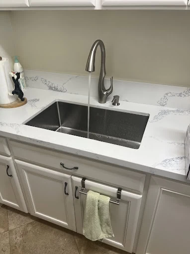 A modern kitchen sink with water running from a silver faucet, set in a white countertop with subtle gray veining. White cabinets below have a towel hanging from a bar. Installed by a plumbing contractor Sacramento County, CA.