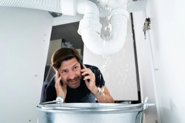 A man on the phone looks worried under a sink as water leaks heavily from a white pipe above a metal bucket, urgently needing plumbing contractor Sacramento County or plumber services El Dorado, CA.