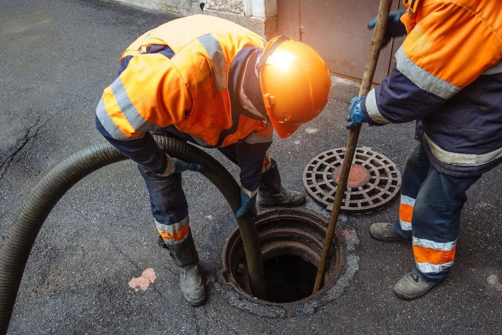 Two workers in orange safety gear and helmets clean a sewer, one holding a large hose and the other using a pole; a manhole cover rests nearby on the asphalt in El Dorado, CA.