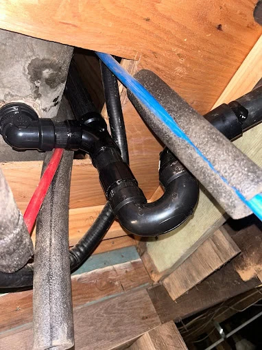 Black plumbing pipes with connectors and insulated water lines (red and blue) run through wooden beams in an unfinished ceiling, possibly in a basement—a common sight for a plumbing contractor Sacramento County or plumber services El Dorado, CA.