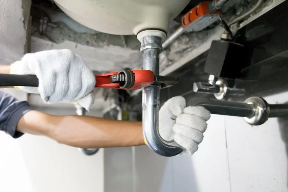 A person wearing white gloves uses a red pipe wrench to tighten or loosen a chrome drain pipe under a sink, performing plumbing maintenance or repair in El Dorado, CA.