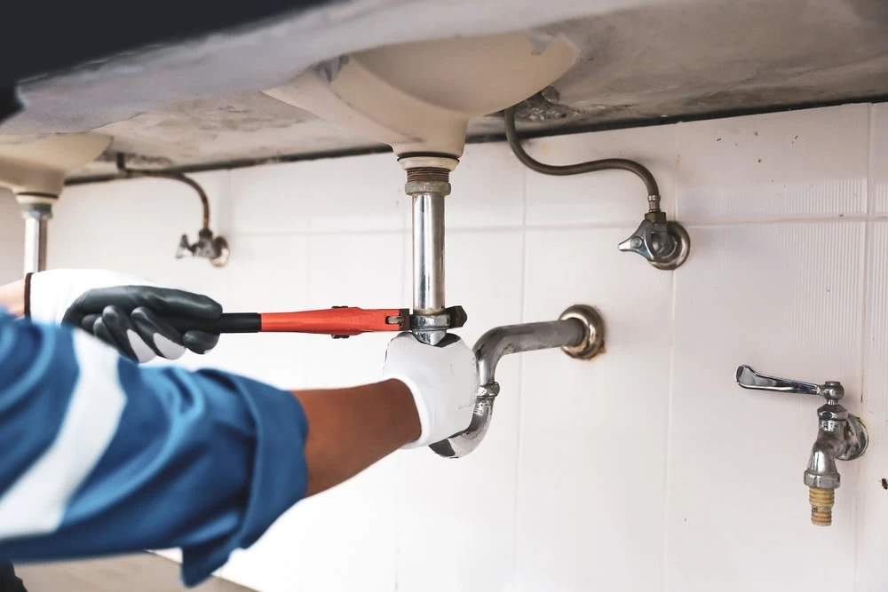 A plumber wearing gloves uses a wrench to tighten or loosen a pipe under a sink, with visible plumbing fixtures and connections against a white tiled wall.