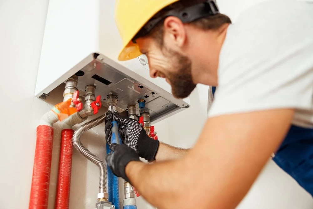 A plumber wearing a yellow hard hat and gloves uses pliers to fix or install pipes connected to a wall-mounted water heater. Red and silver pipes are visible, and he is focused on his work in El Dorado, CA.