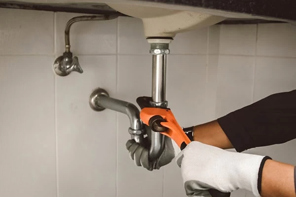 A person wearing gloves uses a wrench to tighten or loosen a pipe under a sink, performing plumbing work in a tiled bathroom or kitchen for a plumbing contractor Sacramento County, CA.