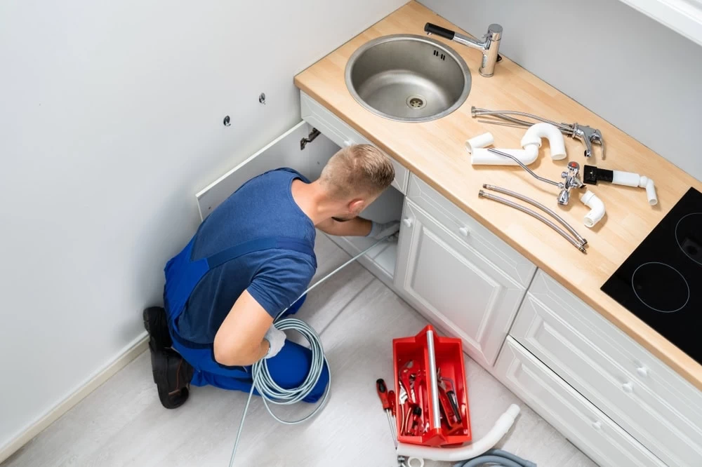 A plumber in blue overalls kneels on the kitchen floor, working under a sink with tools and pipes scattered on the countertop and a red toolbox nearby.