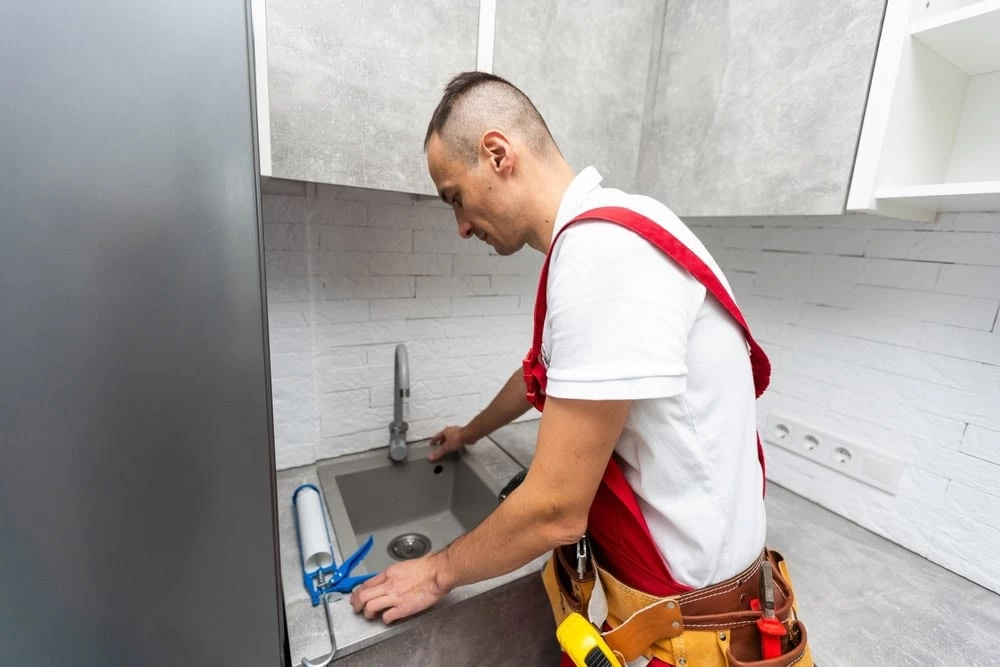 A plumber wearing a tool belt and red suspenders installs or repairs a kitchen faucet in a modern kitchen with gray cabinets and a white brick backsplash in El Dorado, CA.