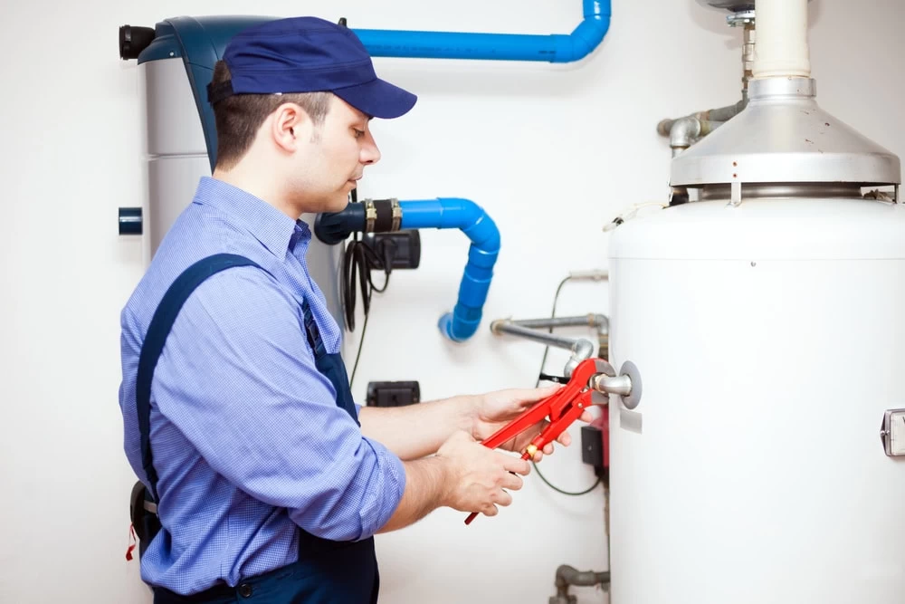 A plumber in a blue uniform and cap uses a large wrench to tighten a fitting on a white water heater in a utility room with blue pipes visible in the background.