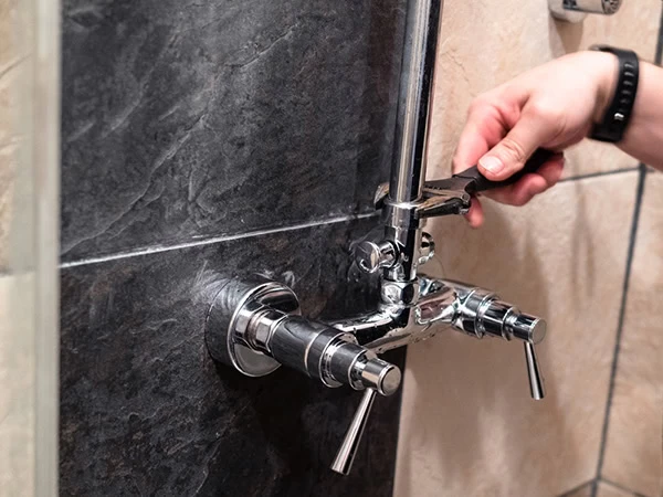 A person’s hand uses a wrench to adjust or repair a chrome shower faucet mounted on a dark tiled bathroom wall, showcasing the expert work offered by plumber services El Dorado, CA.