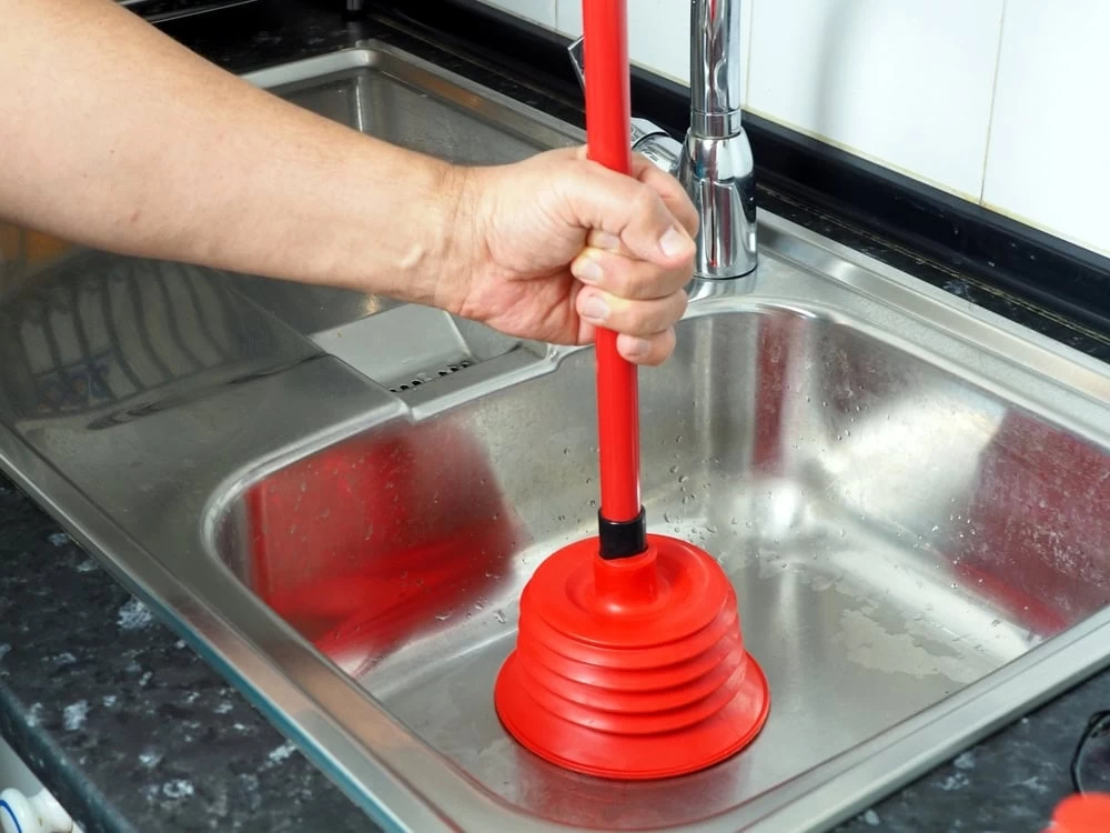 A hand using a red plunger to unclog a steel kitchen sink, with water partially filling the basin and a faucet visible in the background in El Dorado, CA.