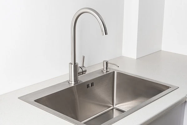 A modern stainless steel kitchen sink with a tall curved faucet and built-in soap dispenser, set in a white countertop against a plain white wall—perfect for those seeking expert plumber services in El Dorado, CA.