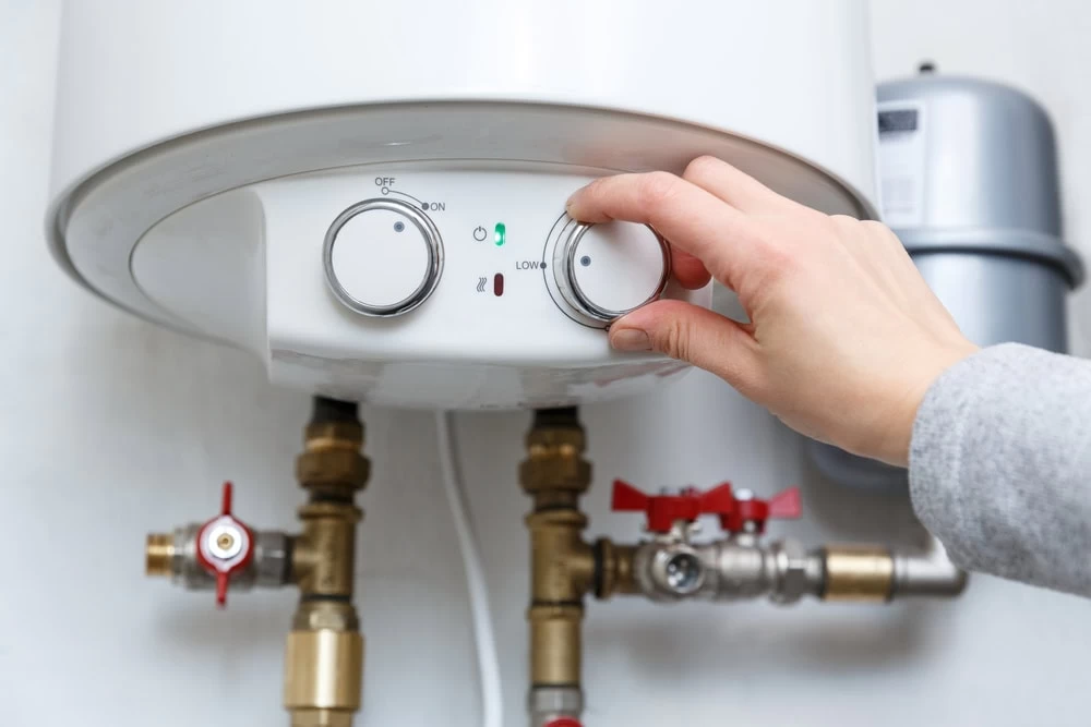 A person’s hand is adjusting the temperature dial on a white electric water heater mounted on a wall, with visible pipes and valves underneath in El Dorado, CA.