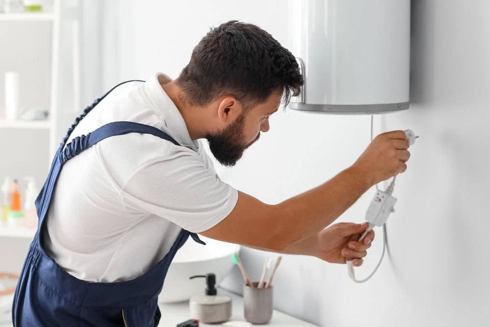 A man in overalls is installing or repairing a water heater mounted on a white wall, inspecting and handling its wires and controls.