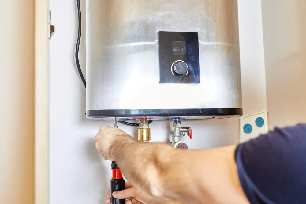 A person uses a screwdriver to repair or install a wall-mounted water heater with a digital display. The individual’s arm is visible, adjusting a fitting on the bottom of the metal unit in El Dorado, CA.