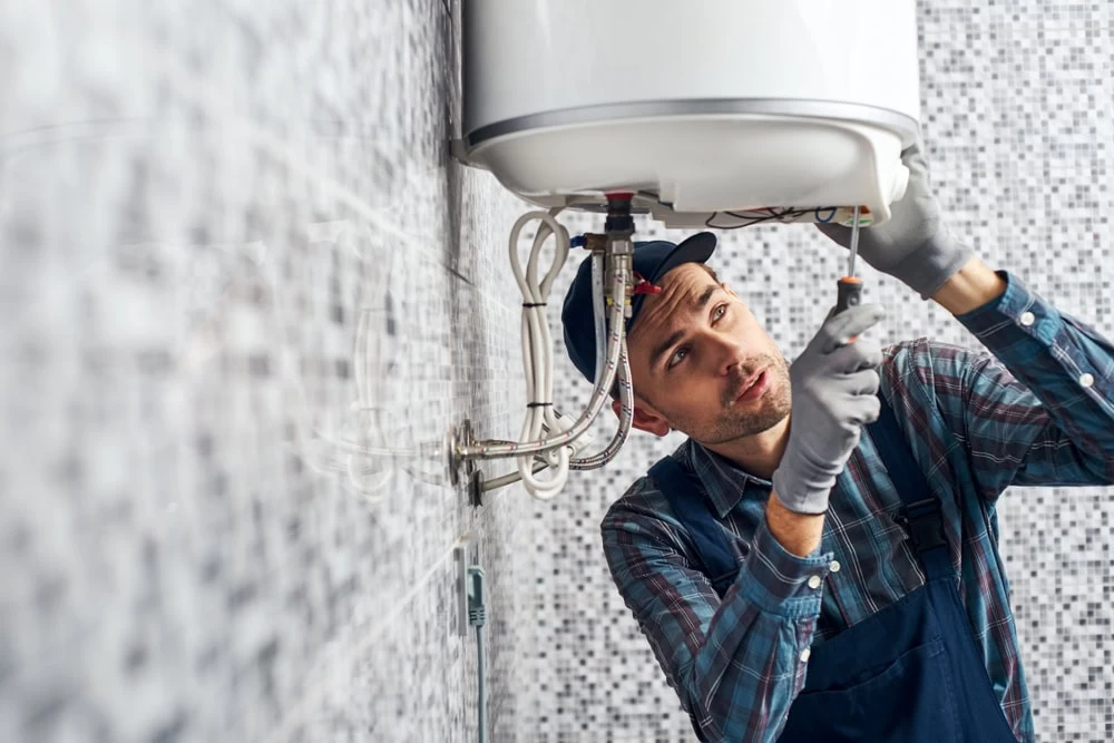 A plumber wearing gloves, a cap, and overalls uses a screwdriver to repair or install a white water heater mounted on a tiled bathroom wall in El Dorado, CA.