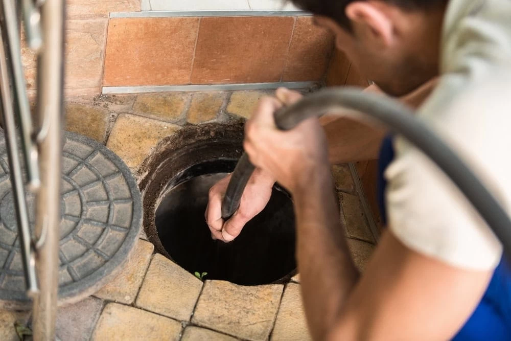 A person wearing a blue uniform uses a hose to clean or unclog a drain or sewer opening on a tiled floor.