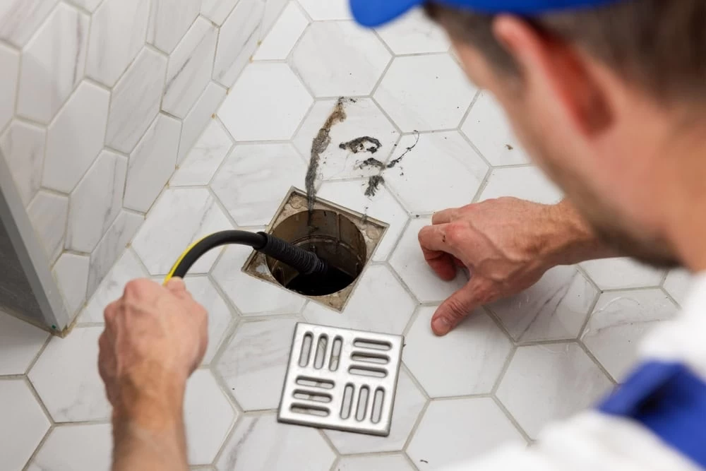 A person uses a tool to clean or inspect a floor drain on a white hexagonal tile surface; the metal drain cover is removed and placed nearby.