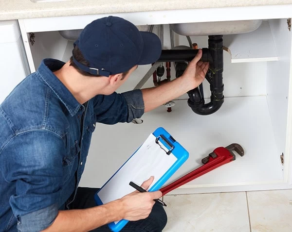 A plumber wearing a blue cap and shirt inspects pipes under a kitchen sink while holding a red pipe wrench and a clipboard with paper and pen.