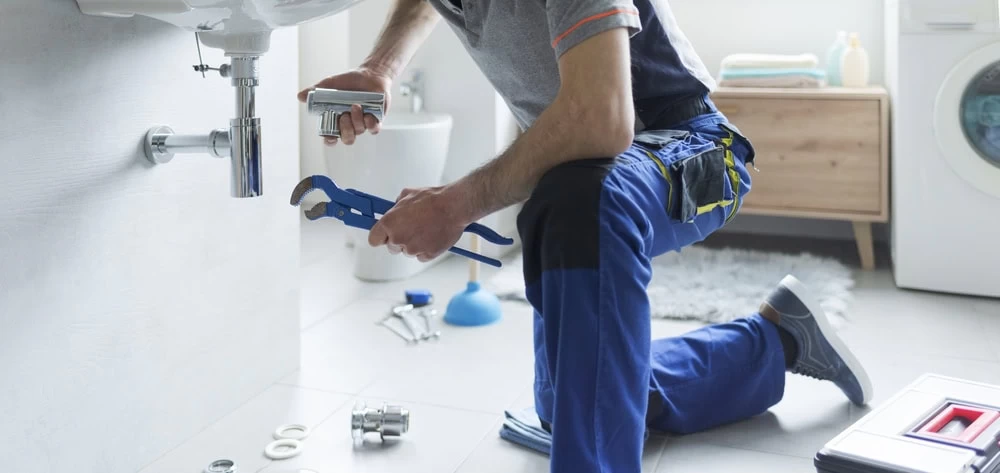A plumber kneels on a bathroom floor, repairing a pipe under a sink with a wrench. Various plumbing tools and parts are scattered nearby, and a washing machine is visible in the background.