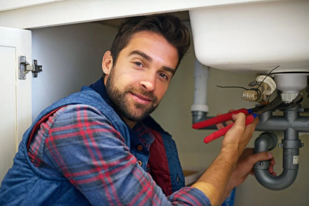 A man wearing a plaid shirt and denim vest is crouched under a sink, using pliers to work on the pipes, and smiling at the camera in El Dorado, CA.