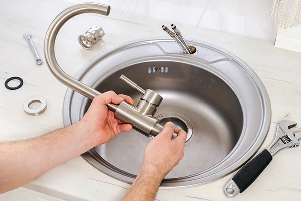 A person installs a new kitchen faucet into a stainless steel sink, with plumbing tools and faucet parts placed nearby on the countertop.